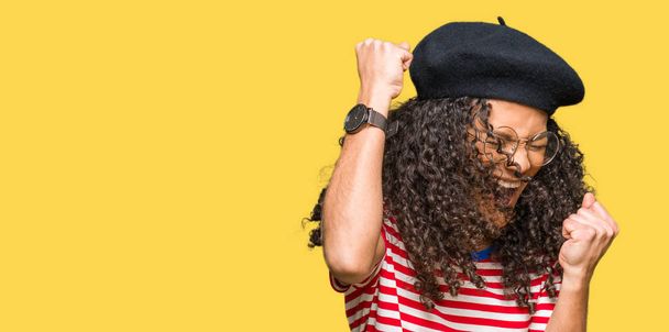 Young beautiful woman with curly hair wearing glasses and fashion beret very happy and excited doing winner gesture with arms raised, smiling and screaming for success. Celebration concept. - Photo, Image