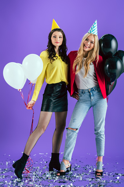 Studio πλάνο της ευτυχισμένος κορίτσια κόμμα καπέλα κρατώντας αερόστατα σε μοβ φόντο - Φωτογραφία, εικόνα