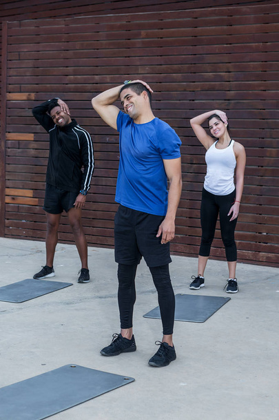 Handsome personal instructor smiling and doing stretching exercise for neck with two multiracial athletes on street - Photo, Image