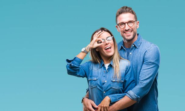 Young couple in love wearing glasses over isolated background doing ok gesture with hand smiling, eye looking through fingers with happy face. - Photo, Image