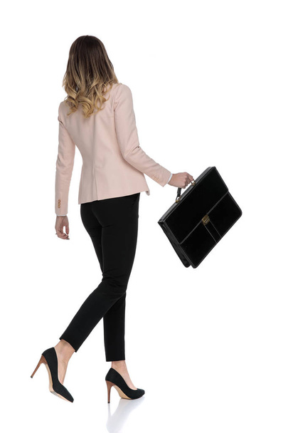 vista trasera de atractiva mujer de negocios caminando hacia un lado sobre fondo blanco y sosteniendo la maleta, imagen de longitud completa
 - Foto, Imagen