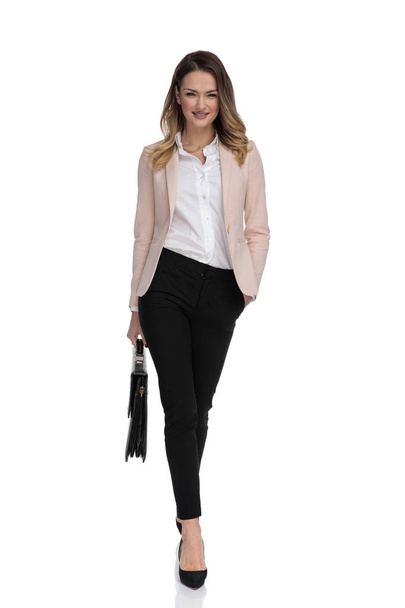 entspannte Geschäftsfrau mit schwarzem Koffer geht auf weißem Hintergrund mit Hand in Tasche zur Arbeit, Ganzkörperbild - Foto, Bild