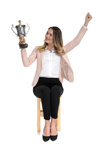 happy businesswoman looks at trophy to side and celebrates with hand in the air while sitting on wooden cahir on white background, full length picture - Photo, Image