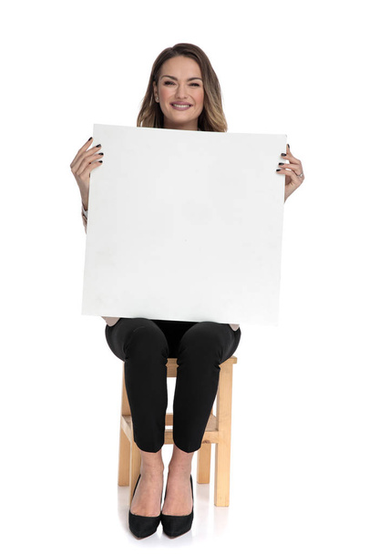 Geschäftsfrau hält leeres weißes Brett, während sie auf einem Holzstuhl auf weißem Hintergrund sitzt, Bild in voller Länge - Foto, Bild