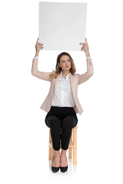 curious smart casual woman holds empty board and looks to side while sitting on wooden stool on white background - Photo, Image