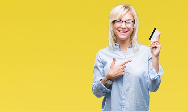 Νέοι επαγγελματίες όμορφη ξανθιά γυναίκα που κρατά πιστωτική κάρτα πέρα από το απομονωμένο υπόβαθρο πολύ χαρούμενος, δείχνοντας με το χέρι και το δάχτυλό - Φωτογραφία, εικόνα