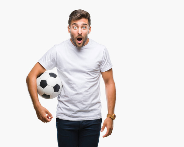 Νέος όμορφος άνθρωπος που κρατά την μπάλα ποδοσφαίρου ποδοσφαίρου πέρα από το απομονωμένο υπόβαθρο φοβάται το σοκ με πρόσωπο έκπληξη, φοβάται και ενθουσιασμένος με φόβο έκφραση - Φωτογραφία, εικόνα
