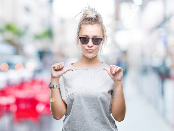 Jeune femme blonde portant des lunettes de soleil sur fond isolé regardant confiant avec le sourire sur le visage, se montrant du doigt fier et heureux
. - Photo, image