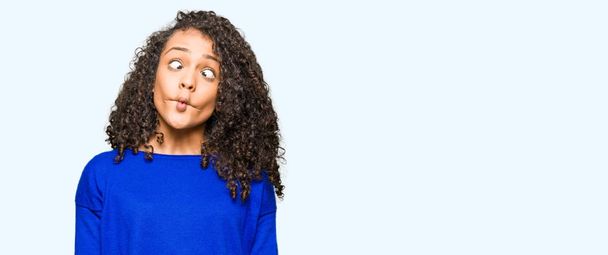 Young beautiful woman with curly hair wearing winter sweater making fish face with lips, crazy and comical gesture. Funny expression. - Foto, afbeelding