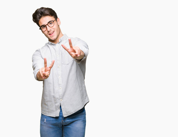 Jonge knappe man met een bril op geïsoleerde achtergrond glimlachend op zoek naar de camera toont vingers doen overwinning teken. Nummer twee. - Foto, afbeelding