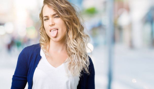 Mooie jonge blonde vrouw op geïsoleerde achtergrond steken tong uit blij met grappige expressie. Emotie concept. - Foto, afbeelding