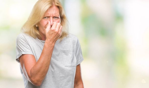 Middle age blonde woman over isolated background smelling something stinky and disgusting, intolerable smell, holding breath with fingers on nose. Bad smells concept. - Photo, Image