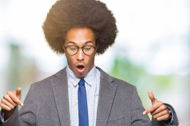 Jonge Afro-Amerikaanse zakenman met het dragen van afro haar bril Pointing omlaag met de vingers laten zien van de advertentie, verbaasd gezicht en open mond - Foto, afbeelding