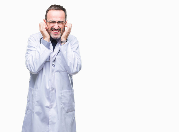 Старший седовласый врач средних лет, одетый в медицинскую униформу, закрывает уши пальцами с раздраженным выражением для шума громкой музыки. Глухая концепция
. - Фото, изображение