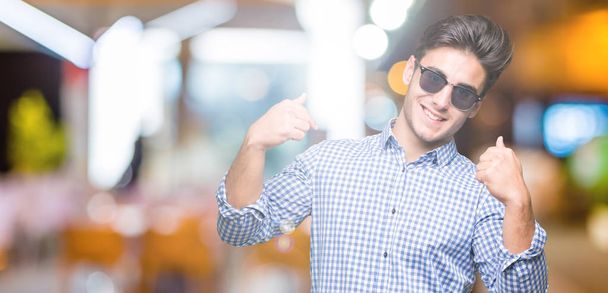 Young handsome man wearing sunglasses over isolated background looking confident with smile on face, pointing oneself with fingers proud and happy. - Photo, Image