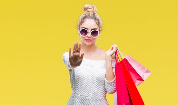 Young beautiful blonde woman holding shopping bags over isolated background with open hand doing stop sign with serious and confident expression, defense gesture - Foto, afbeelding