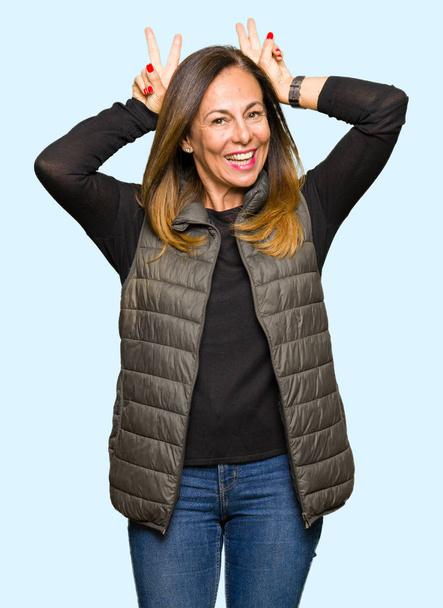 Hermosa mujer de mediana edad con chaleco de invierno Posando divertido y loco con los dedos en la cabeza como orejas de conejo, sonriendo alegre
 - Foto, imagen