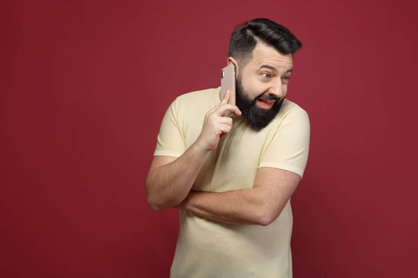 A cute and bearded brunette man on a red background says emotionally on the phone - Фото, зображення