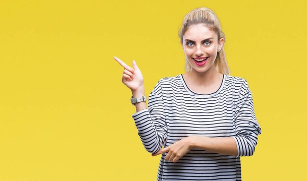 Young beautiful blonde woman wearing stripes sweater over isolated background with a big smile on face, pointing with hand and finger to the side looking at the camera. - Photo, Image