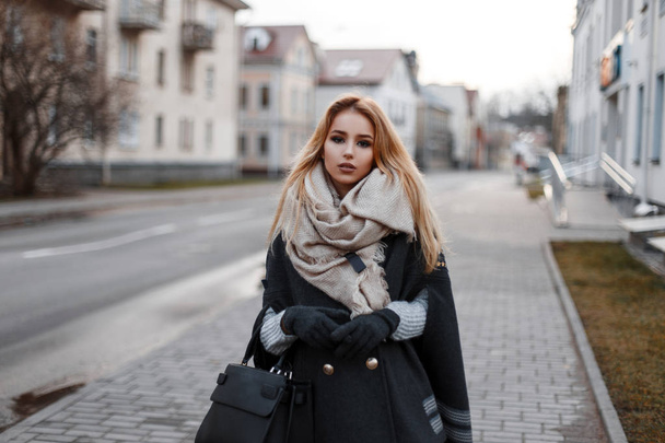 Модна приваблива молода блондинка в вінтажному в'язаному светрі в стильному пальто в теплих рукавичках з вінтажним шарфом з модним чорним шкіряним мішком, що йде по вулиці. Місто мила дівчина
. - Фото, зображення