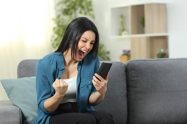 Excited woman reading good news online on smart phone sitting on a couch in the living room at home - Foto, Imagen