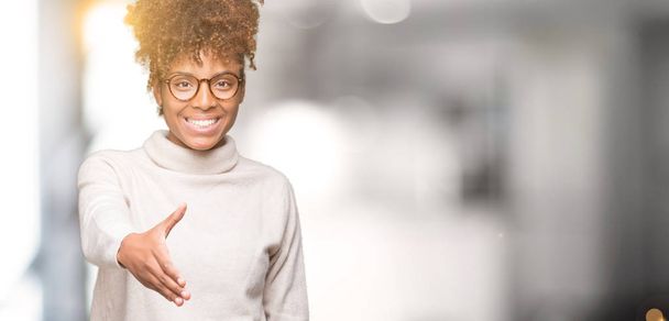 Όμορφη νεαρή αφρικανική αμερικανική γυναίκα που φοράει γυαλιά, πέρα από το απομονωμένο υπόβαθρο χαμογελώντας προσφέροντας φιλική χειραψία ως χαιρετισμό και καλωσόρισμα. Επιτυχημένη επιχείρηση. - Φωτογραφία, εικόνα