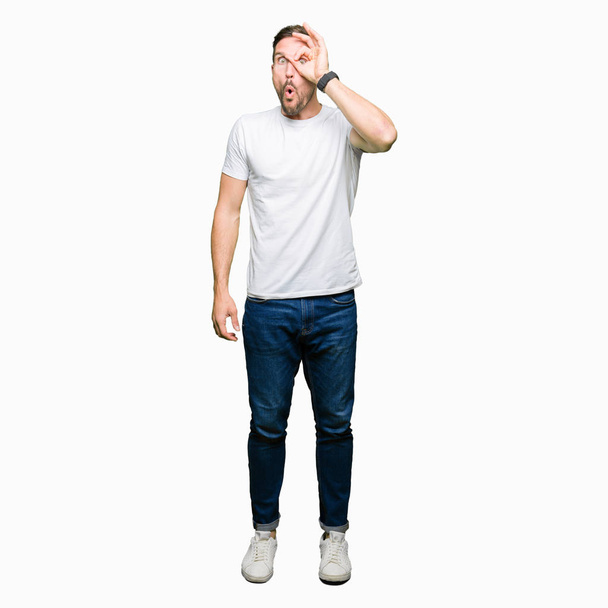 Pohledný muž nosí ležérní bílé tričko dělá ok gesto šokoval s tváří překvapeně, oko dívá skrze prsty. Nevěřící výraz. - Fotografie, Obrázek