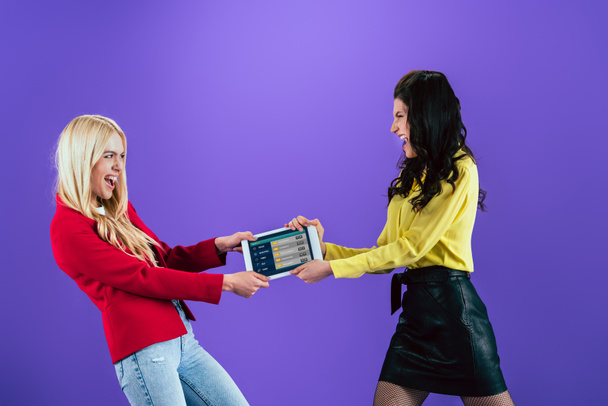 Студийный снимок кричащих девушек, держащих цифровой планшет со спортивным приложением на экране на фиолетовом фоне
 - Фото, изображение