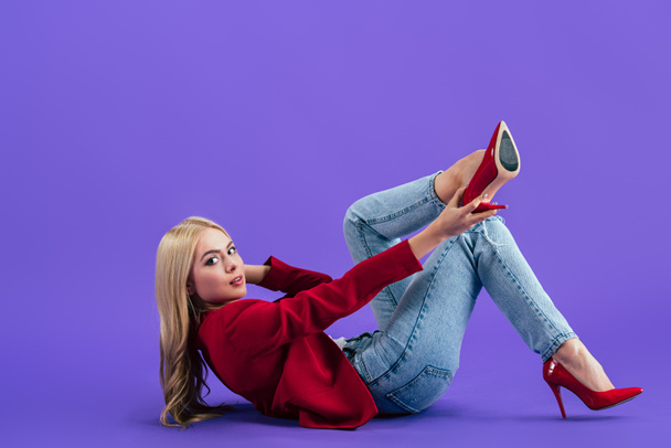 Stylish woman lying on floor and putting on high-heeled shoes on purple background - Foto, Bild