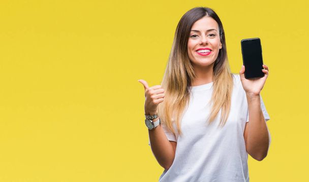 Genç güzel kadın gösteren boş ekran smartphone işaret ve başparmak kadar mutlu yüz gülümseyen tarafı ile gösteren izole arka plan üzerinde - Fotoğraf, Görsel