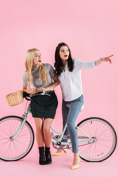 Surprised brunette girl pointing with finger while posing on bicycle with friend on pink background - Photo, Image