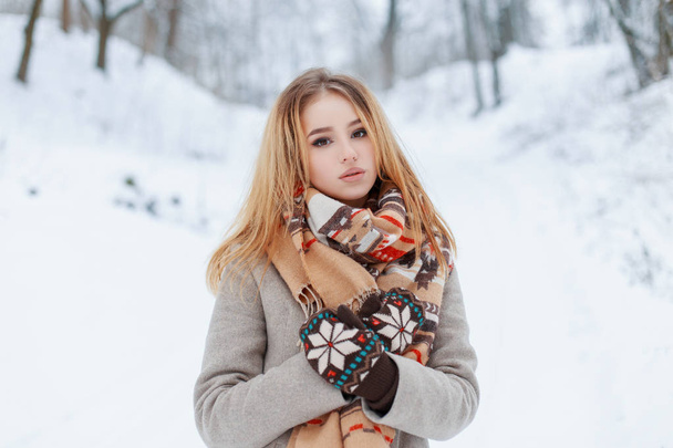 Linda joven encantadora con un elegante abrigo de invierno en guantes vintage de lana con una bufanda beige de lana con un patrón multicolor descansando en la naturaleza en un día frío de invierno. Chica con estilo
. - Foto, imagen
