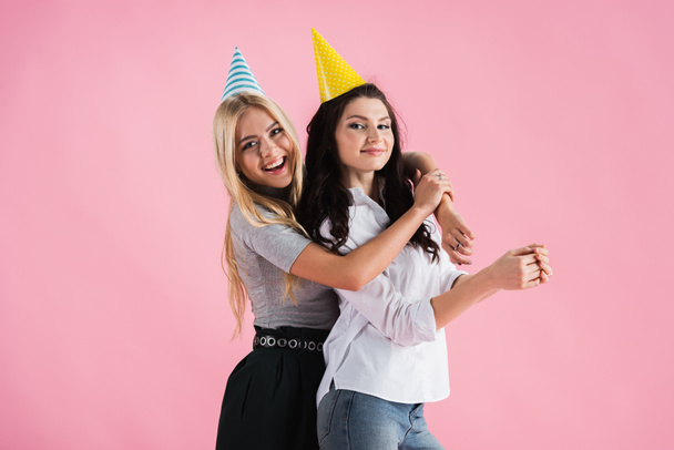 Joyful girls in party hats embracing with smile isolated on pink - Фото, зображення