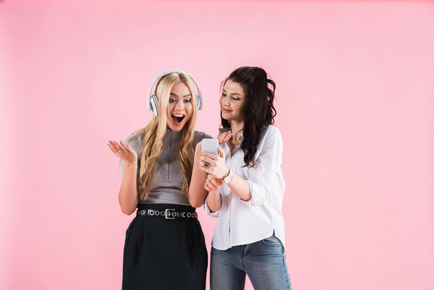 Beautiful excited girls with headphones and smartphone listening music isolated on pink - Photo, image
