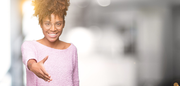 Belle jeune femme afro-américaine portant des lunettes sur fond isolé souriant amical offrant poignée de main comme salutation et accueil. Une entreprise prospère
. - Photo, image