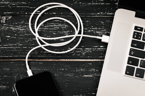 Ένα καλώδιο USB συνδέει το τηλέφωνο και το laptop σας. Στο μαύρο τραπέζι - Φωτογραφία, εικόνα