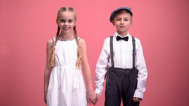 Smiling little boy and girl looking into camera holding hands on pink background - 写真・画像
