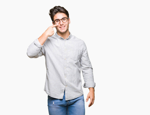 Genç yakışıklı adam gözlük izole üzerinde işaretleme için yüz ve burun, neşeli gülümseyerek el parmak ile arka plan - Fotoğraf, Görsel