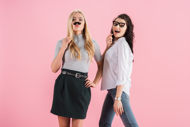 Cheerful girls posing with toy mustache and glasses isolated on pink - Фото, изображение