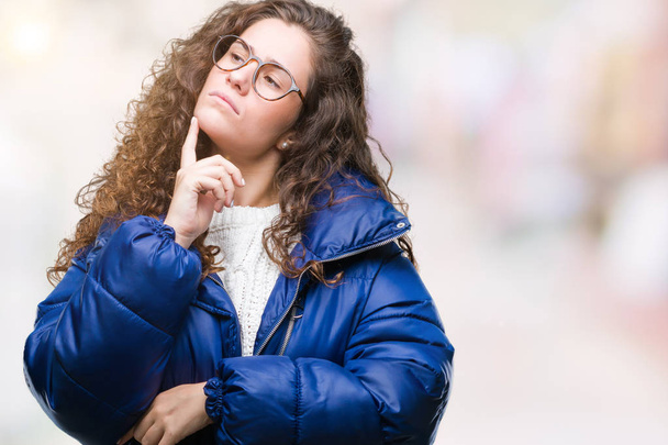 Beautiful young brunette curly hair girl wearing winter coat, glasses and sweater over isolated background with hand on chin thinking about question, pensive expression. Smiling with thoughtful face. Doubt concept. - Photo, Image