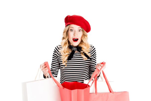 mulher surpreso em boina vermelha olhando para a câmera enquanto segurando sacos de compras isolados no branco
  - Foto, Imagem