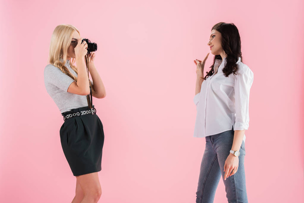 Blondi tyttö ottaa valokuvan, kun brunette tyttö osoittaa rauhan merkki eristetty vaaleanpunainen
 - Valokuva, kuva