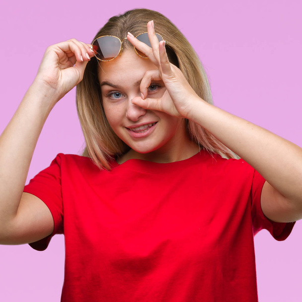 Young caucasian woman wearing sunglasses over isolated background doing ok gesture with hand smiling, eye looking through fingers with happy face. - Fotó, kép