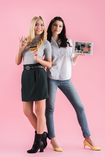 Tytöt osoittavat kunnossa merkki poseeratessaan luottokortilla ja digitaalinen tabletti online-varaus sovellus ruudulla vaaleanpunainen tausta
 - Valokuva, kuva