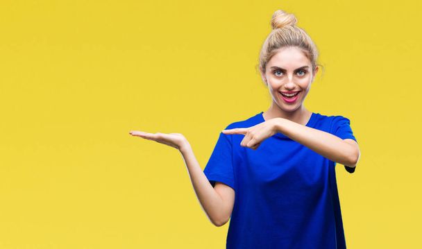 Young beautiful blonde and blue eyes woman wearing blue t-shirt over isolated background amazed and smiling to the camera while presenting with hand and pointing with finger. - Photo, Image