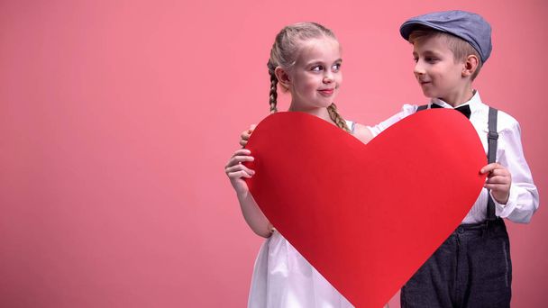 Little romantic kids holding red paper cutout and looking at each other, love - Фото, зображення