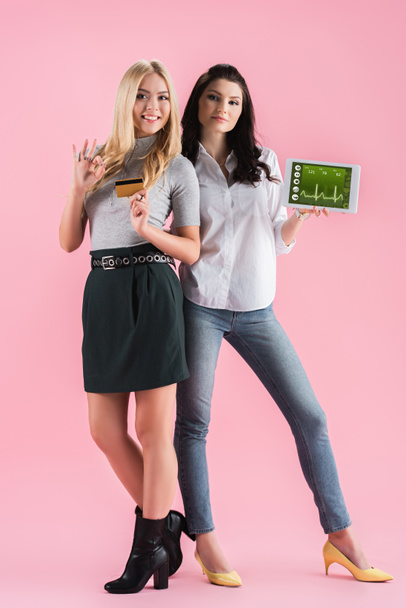 Confident girls holding credit card and digital tablet with fitness app on screen and showing okay sign on pink background - Фото, изображение