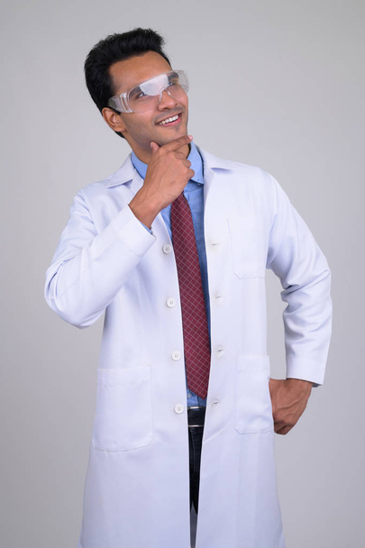 Young happy Indian man doctor with protective glasses thinking - Фото, зображення