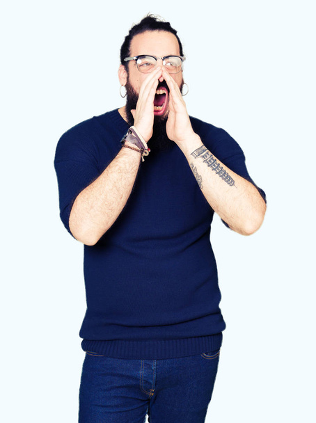 Молодой хипстер с длинными волосами и бородой в очках кричит громко с руками над ртом
 - Фото, изображение