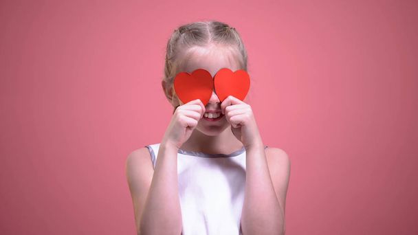 Funny little girl closing eyes with red paper hearts isolated on pink background - Foto, Imagen